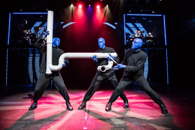 3 men dressed in black with blue skin hold a large pipe